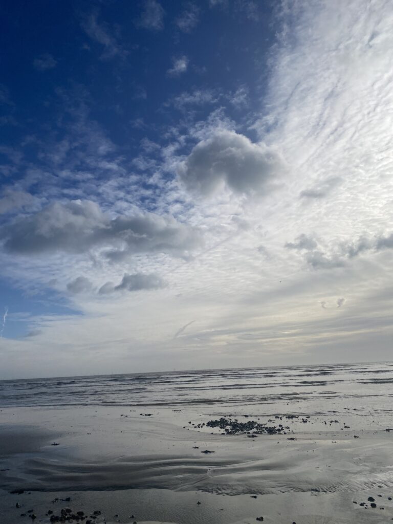 Sandy beach with sand ripples and a few stones. Sea with small waves in the background.Sky is half blue and half filled with clouds that are wispy on the left, going into full cloud on the right and over the sea.