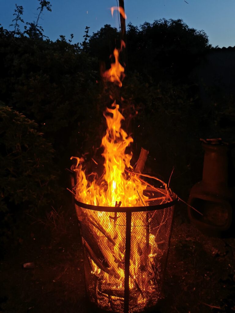 A fire basket filled with burning wood and flaming bright yellow against the black night. Flames lick, high up into the air.