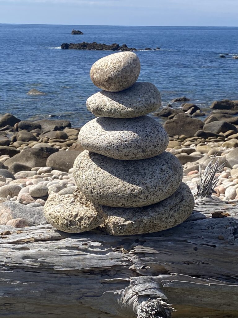 Round pebbles stacked onto of each other with largest at the bottom and smallest on top.  In the background is pebbly beach and the blue sea.